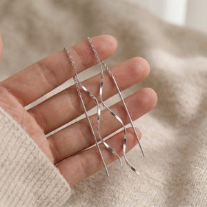 May include: Silver threader earrings with a delicate chain and a wavy, linear design. The earrings have a minimalist style and are shown against a neutral background.