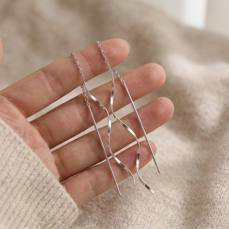 May include: Silver threader earrings with a delicate chain and a wavy, linear design. The earrings have a minimalist style and are shown against a neutral background.