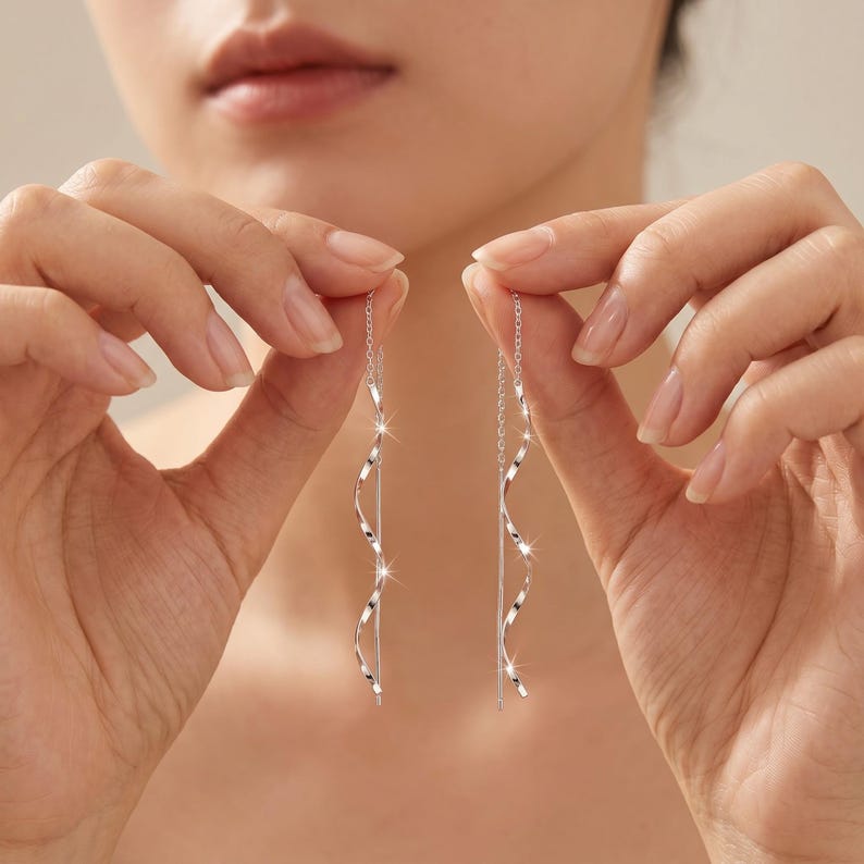 May include: A pair of silver earrings with a spiral design. The earrings are held up by a person's hands. The earrings have a long, thin chain and a curved, twisted design that catches the light.
