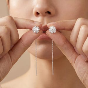 May include: A pair of silver threader earrings featuring a spherical, textured stud component with a radiating, starburst-like design. Each earring consists of a delicate silver chain that extends downward, terminating in a smooth, slender silver bar.
