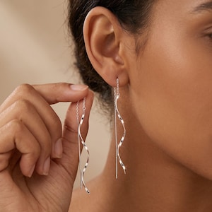 May include: Silver threader earrings with a straight bar and a wavy, spiral design. The earrings are shown in a close-up view, highlighting their delicate and elegant appearance. The earrings are being held up to the ear.