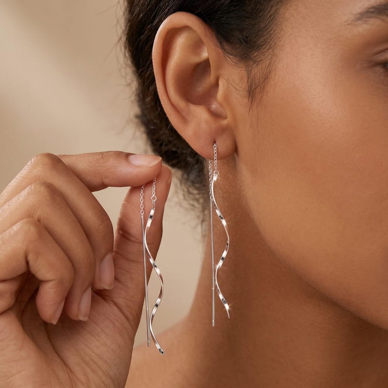 May include: Silver threader earrings with a straight bar and a wavy, spiral design. The earrings are shown in a close-up view, highlighting their delicate and elegant appearance. The earrings are being held up to the ear.