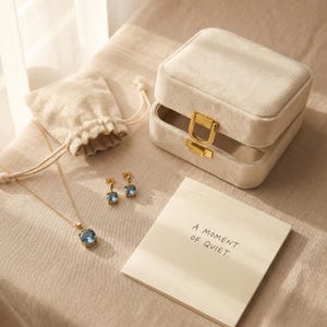 May include: A jewelry set featuring a gold necklace and earrings with blue gemstones, a beige jewelry box, and a small drawstring bag. A notepad with the text "A MOMENT OF QUIET" is also present.