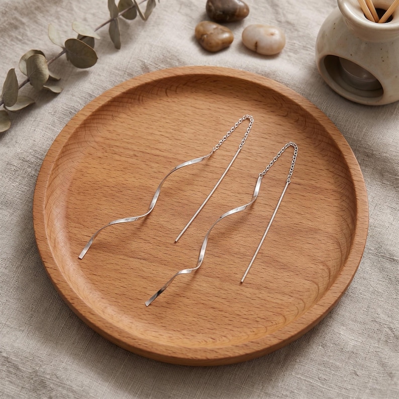 May include: A pair of silver threader earrings with a wavy, textured design. The earrings are displayed on a round, wooden tray. The background includes small stones and a ceramic container.