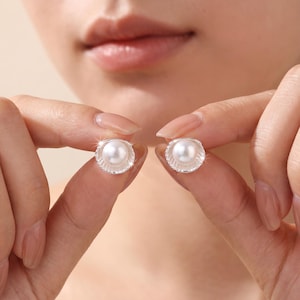 May include: A pair of silver stud earrings, each featuring a white pearl center surrounded by a scalloped, textured border. The earrings are held up close to the camera, showcasing their delicate design and reflective surfaces.