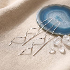 May include: A pair of silver threader earrings with a wavy design. The earrings are displayed on a beige surface with a blue and white geode slice and several clear quartz crystals.