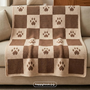 Könnte beinhalten: Eine gehäkelte Decke mit einem Schachbrettmuster aus braunen und beigen Quadraten. Jedes beige Quadrat zeigt einen braunen Pfotenabdruck. Die Decke liegt auf einem beigen Sofa. Der Text "happyhookclub" ist unten rechts zu sehen.