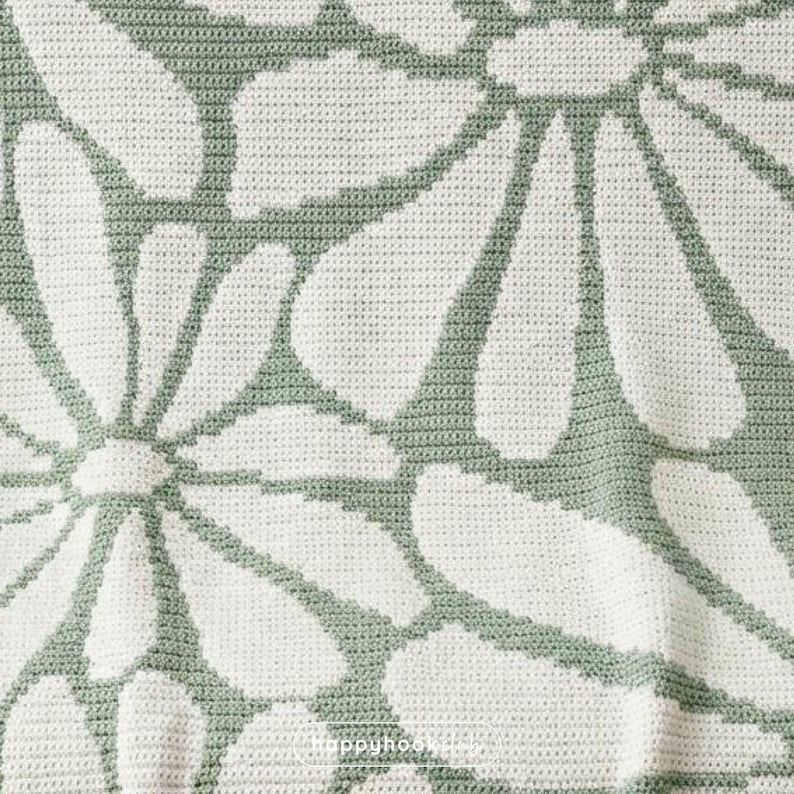 May include: Close-up of a crocheted blanket with a large daisy pattern. The blanket features white daisy petals outlined in a soft green against a white background. The texture of the crochet stitches is visible.