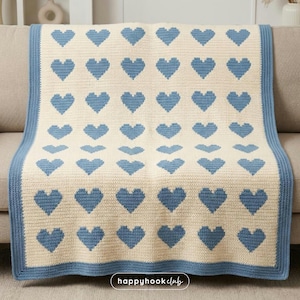 May include: A cream-coloured crocheted blanket with a pattern of blue hearts. The blanket has a blue border and is draped over a sofa. The blanket is handmade and features a repeating heart design.