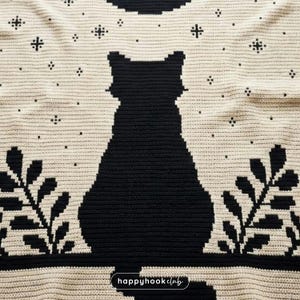 May include: A crocheted blanket featuring a large black cat silhouette against a cream-colored background. The design includes stylized leaves and star-like accents. The blanket has a textured appearance, with the words "happyhookclub" in the bottom right corner.