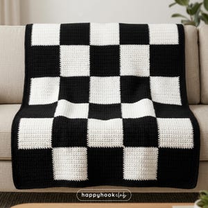 Op de afbeelding: Een zwart-wit geblokte gehaakte deken over een beige bank. De deken heeft een herhalend patroon van grote, vierkante blokken in afwisselende kleuren. De tekst "happyhookclub" is rechtsonder zichtbaar.