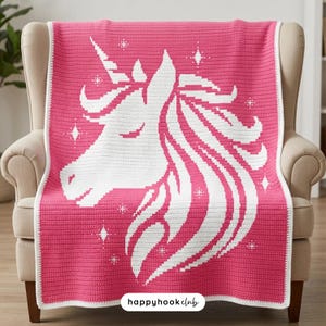 May include: A pink and white crochet blanket with a unicorn design. The blanket features a white border and a large unicorn head in white against a pink backdrop. The blanket is draped over a beige armchair.