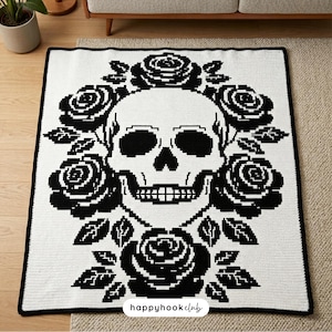 Puede incluir: Una manta de ganchillo blanca y negra con un dise&ntilde;o de calavera rodeada de rosas y hojas negras. La manta tiene un borde negro y el texto "happyhookclub" es visible en la parte inferior.