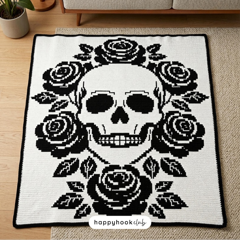 Puede incluir: Una manta de ganchillo blanca y negra con un dise&ntilde;o de calavera rodeada de rosas y hojas negras. La manta tiene un borde negro y el texto "happyhookclub" es visible en la parte inferior.