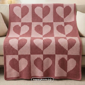 Puede incluir: Una manta de ganchillo hecha a mano en tonos rosa y rosa. La manta presenta un patrón repetido de corazones dentro de un diseño a cuadros. La manta está colocada sobre un sofá beige. El texto "happyhookclub" es visible en la esquina inferior derecha.