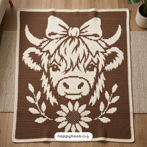Puede incluir: Una manta de ganchillo marrón y crema con un diseño de vaca de las Tierras Altas. La cara de la vaca es crema, con un lazo, cuernos y un girasol. La manta tiene un borde crema y el texto "happyhookclub".