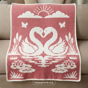 以下が含まれることがあります： ピンクと白のかぎ針編みのブランケット。白の縁取りがあり、2羽の白鳥がハート型を形成し、太陽、雲、蝶、スイレンが描かれています。「happyhookclub」の文字が右下にあります。