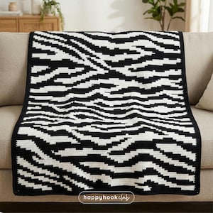 Pode incluir: Uma manta de crochê preta e branca com um padrão de listras de zebra. A manta está sobre um sofá bege. A manta tem uma borda preta e as palavras "happyhookclub" são visíveis no canto inferior direito.