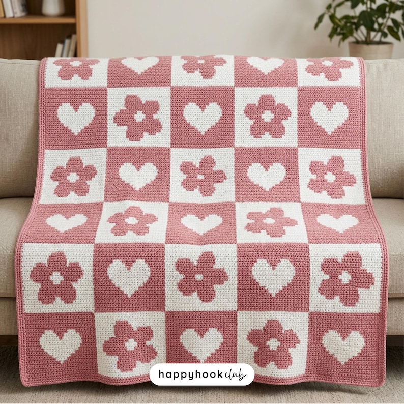 May include: A crocheted blanket with a checkerboard pattern of pink squares with white hearts and flowers. The blanket is draped over a beige sofa. The text "happyhookclub" is in the bottom right corner.