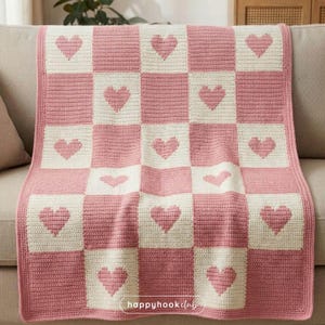 Könnte beinhalten: Eine rosa-weiß gehäkelte Decke mit Schachbrettmuster. Jedes Quadrat zeigt ein Herzdesign. Die Decke ist über ein Sofa drapiert. Der Text "happyhookclub" ist unten rechts sichtbar.