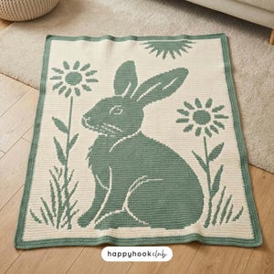 Puede incluir: Una manta de ganchillo rectangular que presenta un conejo de color verde salvia sentado en el centro, flanqueado por dos flores estilizadas y un motivo de sol arriba, sobre un fondo crema con un borde verde salvia y una etiqueta de logo.