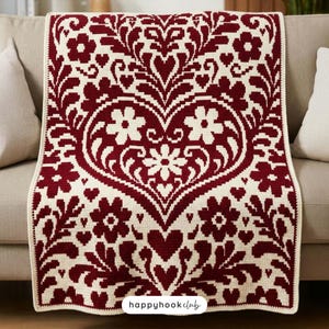 Puede incluir: Una manta de ganchillo rectangular con un patrón simétrico en rojo oscuro y crema. El diseño muestra un gran corazón central rodeado de intrincados motivos florales, hojas y formas de corazón más pequeñas. El texto "happyhookclub" es visible.