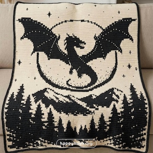 Könnte beinhalten: Gehäkelte Decke mit schwarzem Drachendesign auf cremefarbenem Hintergrund. Das Design zeigt einen Mond, Berge und Bäume. Die Decke hat einen schwarzen Rand und den Text "happyhook club".
