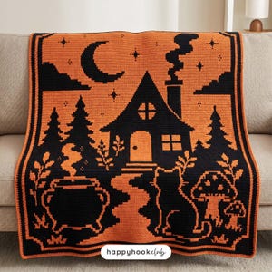 Puede incluir: Una manta de ganchillo rectangular con un diseño en negro y naranja. El patrón muestra una casa con chimenea humeante, una luna creciente, estrellas, pinos, un caldero humeante, un gato y setas, junto con el texto happyhookclub.