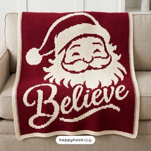 Op de afbeelding: Een donkerrode en crèmekleurige gehaakte deken met een grote afbeelding van de Kerstman met een muts en het woord Believe in cursief schrift onder het gezicht. De deken heeft een crèmekleurige rand en een klein wit label aan de onderkant.
