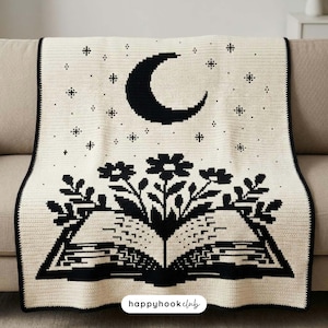 Op de afbeelding: Een crèmekleurige gehaakte deken met een zwart grafisch ontwerp van een open boek waar bloemen uit groeien, bekroond door een halve maan en sterren. De tekst "happyhookclub" staat in het zwart gedrukt op een wit label aan de onderkant.