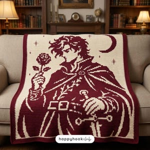 May include: A cream and deep red crochet blanket featuring a graphic design of a person with messy hair wearing a cloak, holding a rose in one hand and a sword in the other, with a crescent moon and stars in the background. The text happyhookclub is visible.