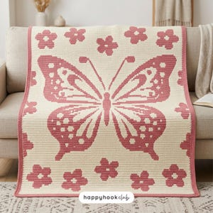 Könnte beinhalten: Eine rechteckige Häkeldecke mit einem großen zentralen Schmetterlingsmotiv und kleineren Blumenmustern in Altrosa auf cremefarbenem Hintergrund. Die Decke hat einen einfarbigen rosa Rand und enthält den Text happyhookclub am unteren Rand.