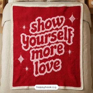 May include: A red crocheted blanket with the words "show yourself more love" in white. The blanket has a white border and starburst designs. The blanket is displayed on a bed.