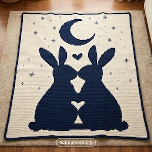 Puede incluir: Una manta de ganchillo color crema con un diseño azul marino de dos conejos besándose bajo una luna creciente y estrellas. La manta tiene un borde azul marino y está colocada sobre una alfombra de tonos neutros. La manta está hecha a mano.