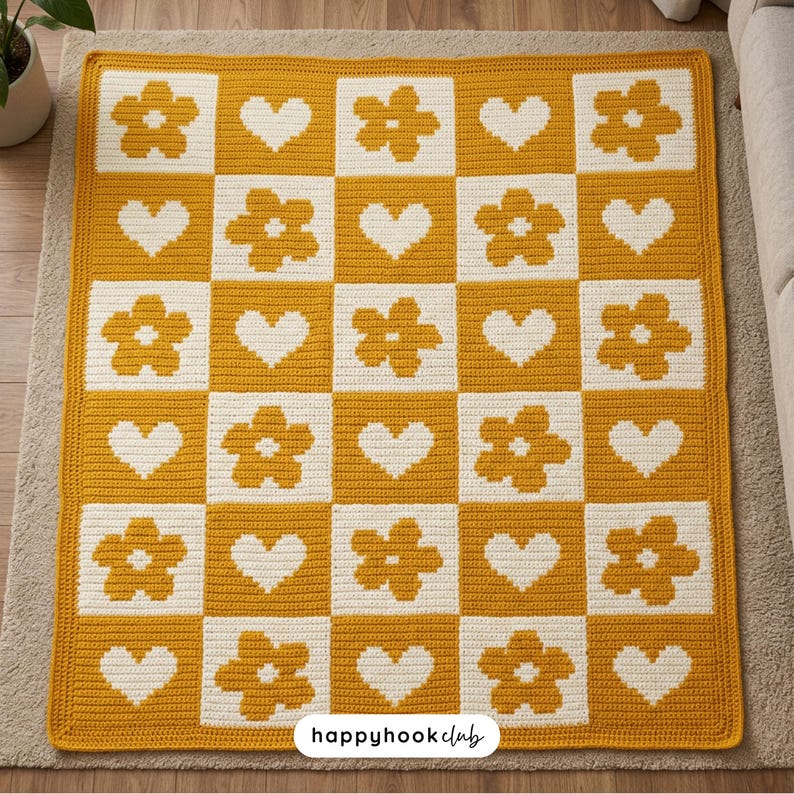 May include: A handmade crochet blanket in a checkerboard pattern. Alternating squares feature a mustard yellow background with white flowers and hearts. The blanket is made of soft yarn and has the text "happyhookclub" at the bottom.
