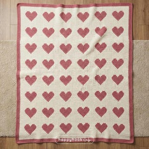 May include: A cream-colored crocheted blanket featuring a pattern of pixelated pink hearts. The blanket has a pink border and the text "happyhookclub" in the bottom right corner. This handmade blanket is perfect for home decor.