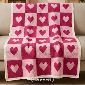 Op de afbeelding: Een handgemaakte gehaakte deken met een dambordpatroon van roze en fuchsia vierkanten, elk met een hartmotief. De deken heeft een crèmekleurige rand en is over een bank gedrapeerd. De tekst "happyhook club" is rechtsonder zichtbaar.