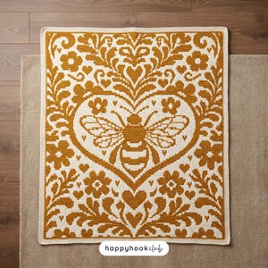 May include: A rectangular crochet blanket featuring a mustard yellow and cream color scheme. The design displays a central bee motif enclosed within a heart shape, surrounded by intricate floral and leaf patterns. The text happyhookclub is visible.