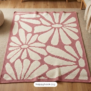 May include: A crocheted blanket with a large floral design. The blanket features a pattern of white daisy-like flowers against a pink background. The text "happyhookclub" is in the bottom right corner. The blanket is approximately 60 inches by 70 inches.