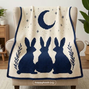 Può includere: Coperta all'uncinetto color crema con bordo blu navy, con tre sagome di conigli, una luna crescente e motivi a stella. La coperta è drappeggiata su una sedia, mostrando un design accogliente e fatto a mano. Il testo "happyhook club" è visibile.