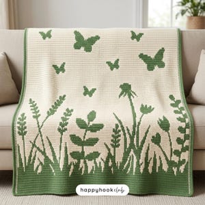 May include: A cream-colored crocheted blanket with a green butterfly and floral design. The blanket features a border of green grass and plants. The text "happyhookclub" is visible in the lower right corner.