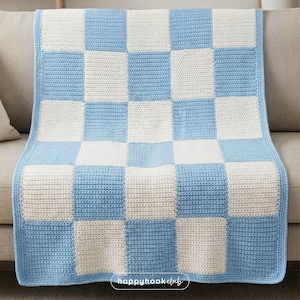Può includere: Una coperta all'uncinetto con un motivo a scacchiera in azzurro e bianco. La coperta è drappeggiata su un divano, mostrando la sua consistenza e il suo design. Il bordo della coperta è azzurro e i quadrati misurano circa 25 cm per 25 cm.