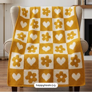 May include: A mustard yellow and cream-colored crocheted blanket with a checkerboard pattern of flowers and hearts. The blanket is draped over a beige armchair. The text "happyhookclub" is visible in the bottom right corner.