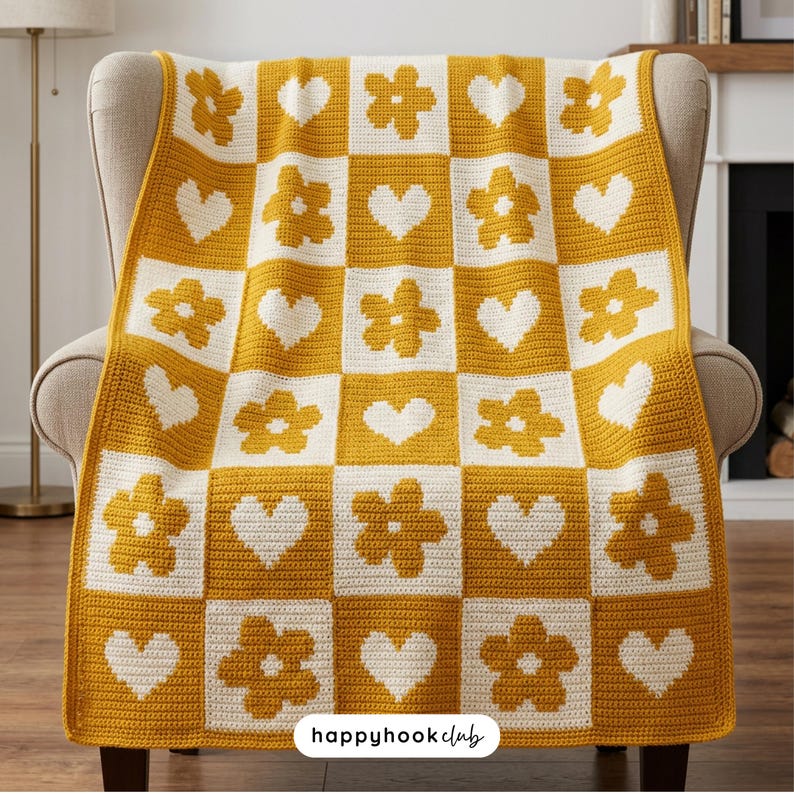 May include: A mustard yellow and cream-colored crocheted blanket with a checkerboard pattern of flowers and hearts. The blanket is draped over a beige armchair. The text "happyhookclub" is visible in the bottom right corner.