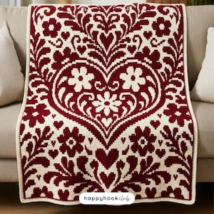 Può includere: Una coperta all'uncinetto bordeaux e crema con un grande motivo floreale a forma di cuore. La coperta presenta un motivo ripetuto di fiori, cuori e foglie. La coperta è drappeggiata su un divano.