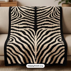 Könnte beinhalten: Eine gehäkelte Decke in Schwarz und Beige mit einem Zebrastreifenmuster. Die Decke ist über ein beiges Sofa drapiert. Die Decke hat einen schwarzen Rand und den Text "happyhookclub" in der unteren rechten Ecke.