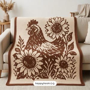 Può includere: Una coperta rettangolare all'uncinetto con un motivo bicolore marrone e crema. Il design mostra una gallina centrale circondata da tre grandi girasoli e fogliame. La coperta ha un bordo marrone solido e il testo happyhookclub.