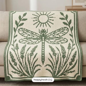 Könnte beinhalten: Eine cremefarbene Häkeldecke mit einem dunkelgrünen Grafikdesign einer großen Libelle unter einer Sonne. Das Muster umfasst flankierende Blattzweige und hohes Gras, alles umschlossen von einem rechteckigen Rand. Der Text lautet happyhookclub.