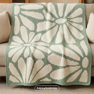 Può includere: Una coperta all'uncinetto con un grande motivo floreale bianco e verde salvia. La coperta è drappeggiata su un divano, mostrando la lavorazione dettagliata e il contrasto cromatico. Il testo "happyhook club" è in basso a destra.