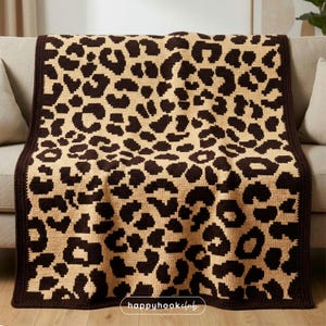 May include: A tan and brown crochet blanket featuring a leopard print pattern. The blanket is draped over a beige sofa. It has a dark brown border. The text "happyhookclub" is visible in the bottom right corner of the blanket.